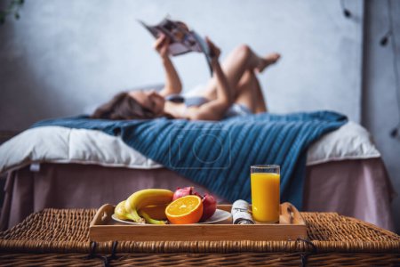 Photo for Beautiful young girl is reading while lying on bed at home, a tray with fruits in the foreground in focus - Royalty Free Image