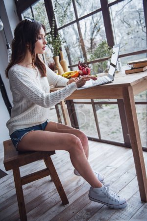 Foto de Hermosa chica joven en ropa casual está trabajando con un ordenador portátil mientras está sentado en la mesa de madera en casa - Imagen libre de derechos
