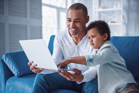 Foto de Feliz padre e hijo en ropa casual están utilizando un ordenador portátil mientras está sentado en el sofá en casa, papá está sonriendo - Imagen libre de derechos