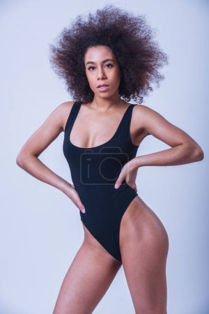 Beautiful woman in black bodysuit is posing at camera akimbo, on a grey background