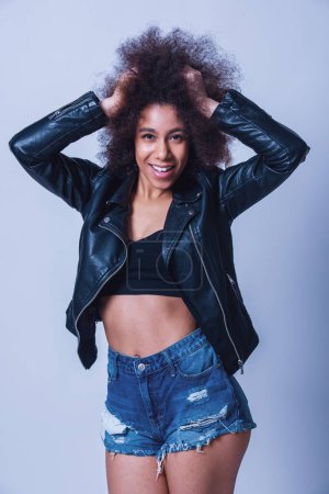 Photo for Beautiful woman in black top, jeans shorts and leather jacket is playing with her hair and smiling, on a grey background - Royalty Free Image
