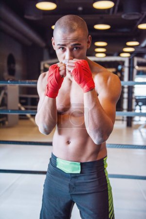 Téléchargez les photos : Beau jeune boxeur, ses mains enveloppées dans un bandage rouge, est debout dans la pose de boxeur dans la salle de gym et en regardant la caméra - en image libre de droit