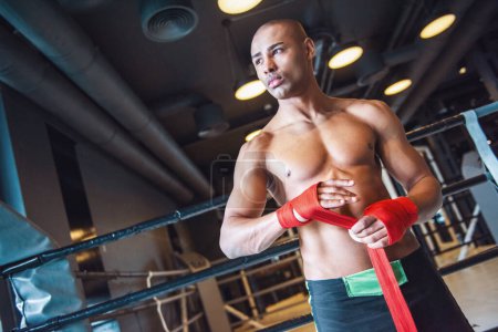 Téléchargez les photos : Beau jeune boxeur emballe les mains avec un bandage rouge et regarde loin tout en se préparant à s'entraîner dans la salle de gym - en image libre de droit