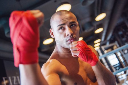 Téléchargez les photos : Beau jeune boxeur, ses mains enveloppées dans un bandage rouge, est debout dans la pose de boxeur dans la salle de gym - en image libre de droit