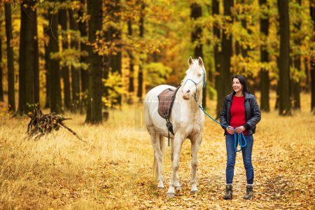 Foto de Mujer joven y un caballo blanco, que atraviesa un sendero forestal en el colorido otoño - Imagen libre de derechos