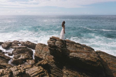 Téléchargez les photos : Vue aérienne de drone de mariée romantique, fille brune en robe de mariée blanche avec les épaules ouvertes posant avec la mer et les roches en arrière-plan. Jeune femme élégante debout sur le bord des falaises et regardant l'océan - en image libre de droit