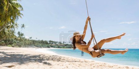 Téléchargez les photos : Modèle de fille de plage bikini en maillot de bain noir s'amusant sur balançoire sur le bungee de palmier sur le rivage de la mer tropicale pendant les vacances d'été. Photo de bannière de haute qualité avec espace de copie pour le texte. - en image libre de droit