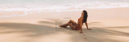 Foto de Bikini mujer de playa en traje de baño rojo descansando bajo la sombra de la palmera en la orilla del mar tropical con arena blanca mirando hacia otro lado. Vista lateral del modelo de ajuste magnífico durante las vacaciones de verano. Banner foto de alta calidad - Imagen libre de derechos