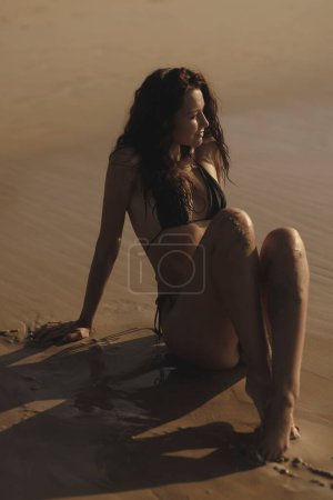 Foto de Summer is the perfect time to be sexy. Fashion shot of a gorgeous young adult woman model in a trendy black bikini sitting in an erotic pose on the beach during her vacation. High quality photo - Imagen libre de derechos