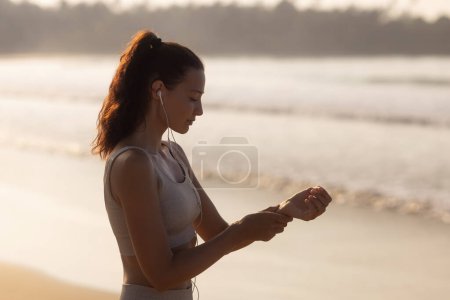 Téléchargez les photos : Silhouette of healthy sports woman in fitness clothes measures pulse by hand on the beach in the morning . High quality photo - en image libre de droit