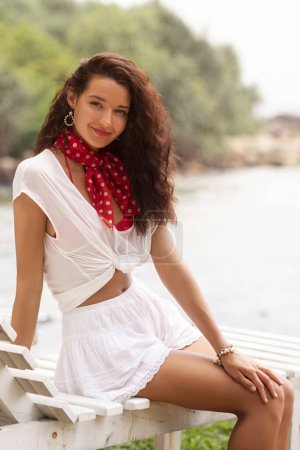 Téléchargez les photos : Superbe belle jeune fille caucasienne assise sur le lit sur une plage vêtue d'une robe blanche élégante, souriant en regardant la caméra - en image libre de droit