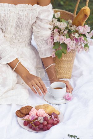 Téléchargez les photos : Une belle mariée avec une élégante tasse de thé profite d'un pique-nique romantique sur une couverture vintage, entourée de détails rustiques et de friandises savoureuses comme les fruits, les biscuits et les croissants. - en image libre de droit