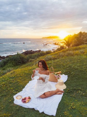Foto de Una mujer joven adulta en un vestido de novia vintage disfruta de un picnic romántico en la playa al atardecer. - Imagen libre de derechos