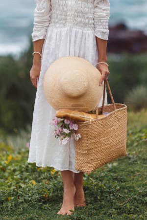 Téléchargez les photos : Une fille à la mode, boho se tient pieds nus dans une robe blanche chic tenant un bouquet créatif de fleurs et un sac de plage en osier rétro dans ses mains, prêt pour un pique-nique pendant les vacances - en image libre de droit