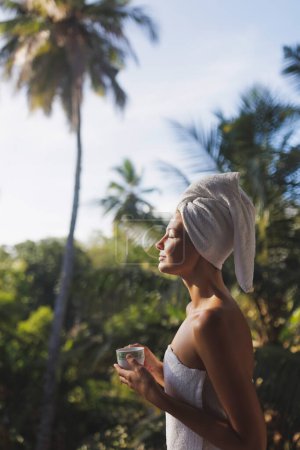 Photo for Good morning. Close-up portrait of a happy woman with a cup of coffee or tea in a white towel after showering, standing on an open resort balcony against jungles with palm trees during a perfect - Royalty Free Image