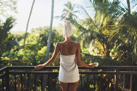 Foto de Buenos días, naturaleza. Vista trasera de una mujer feliz con una taza de té en una toalla blanca después de una ducha, de pie en un balcón abierto contra las palmeras durante una mañana perfecta en un país tropical - Imagen libre de derechos