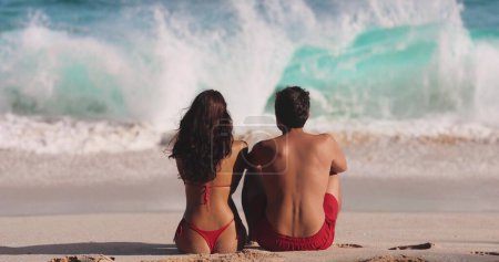 Foto de Hombre y mujer hablando mientras están sentados junto al océano en una playa de arena. Una mujer caucásica morena y un hombre están sentados de espaldas a la cámara, mirando las olas. El concepto de un romántico - Imagen libre de derechos