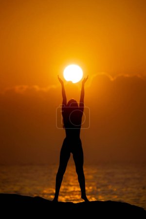  Ganzkörpersilhouette einer jungen sportlichen Frau, die eine Sonnenscheibe in den Händen hält. Konzept für Freiheit und Spiritualität. 