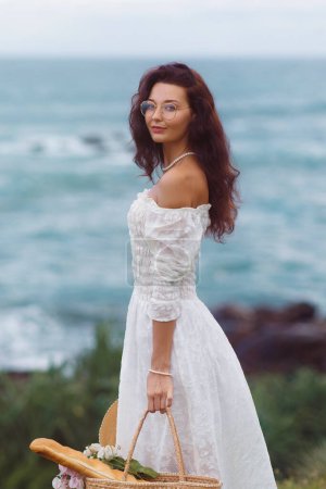 Photo for Stylish summer beach outfit for a boho picnic by the sea of romantic women. Adorable chic standing in a fashionable boho-style white dress with vintage accessories and a flower bouquet in a beach bag - Royalty Free Image