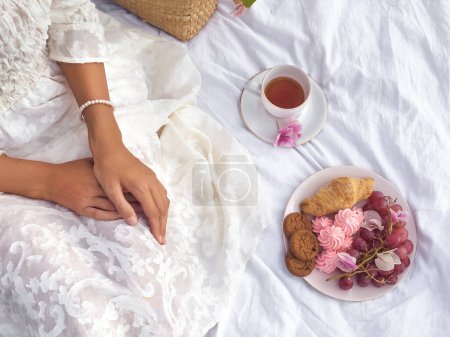 Photo for A beautiful bride with a stylish bun enjoys a romantic picnic on a vintage blanket, surrounded by rustic details and tasty treats like fruit, cookies, mugs of tea, and croissants. - Royalty Free Image