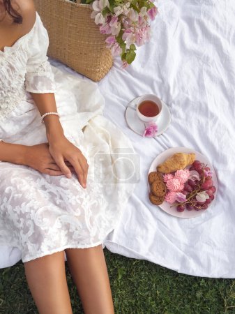 Photo for A young woman sits stylishly on a picnic blanket in an outdoor park, enjoying a romantic and rustic meal of cookies, croissants, a cup of tea, and grapes. - Royalty Free Image