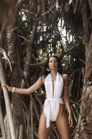 Téléchargez les photos : Prise de vue mode d'un modèle de plage multiracial femme de luxe avec un maquillage et une coiffure parfaits dans des maillots de bain modernes blancs élégants en forêt tropicale - en image libre de droit