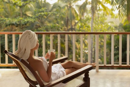 Photo for Portrait of a happy woman with a cup of coffee or tea in a white towel after showering, standing on an open resort balcony against a nature background during a perfect morning in a tropical country - Royalty Free Image