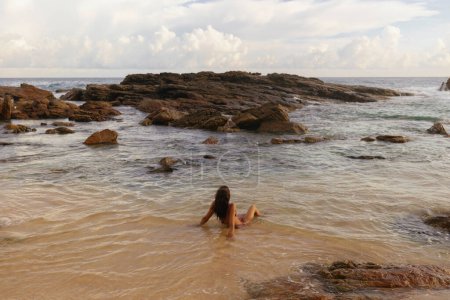 Téléchargez les photos : Bikini plage femme en maillot de bain rouge baignade dans l'eau de mer à la côte rocheuse et profite de l'été. Vue arrière. Vacances de rêve et week-end vacances d'été. Photo de haute qualité - en image libre de droit