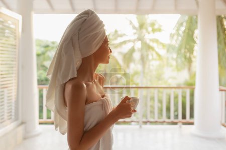 Photo for Perfect sunny morning of beautiful girl in tropical country. Side view of a happy woman with a cup of coffee or tea in a white towel after shower, standing on an open terrace during a perfect morning - Royalty Free Image
