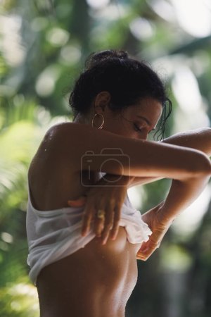 Téléchargez les photos : Jeune femme sexy se déshabillant à l'extérieur sur fond tropical déconcentré, enlevant son T-shirt humide. Jeunesse et séduction. Photo de haute qualité - en image libre de droit