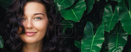 Foto de Belleza natural retrato de cerca de la joven morena feliz modelo femenino con la piel perfecta, maquillaje natural y cabello brillante saludable en frente de la selva entre las plantas exóticas. Cuidado de la piel de alta calidad y - Imagen libre de derechos