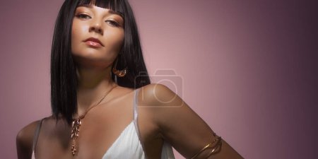Photo for Queen Cleopatra studio beauty portrait. Young adult fashion woman with perfect makeup and stylish straight bob haircut wig, wearing fashionable golden jewelry over pink background. High quality - Royalty Free Image