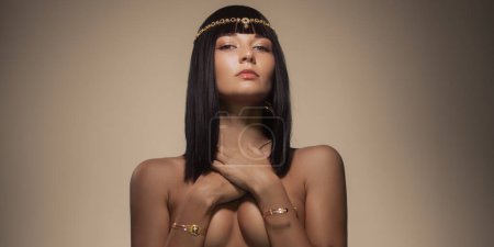 Téléchargez les photos : Beauté mode portrait de femme nue reine style cleopatra couvrant ses seins par les mains, en regardant la caméra, posant nue avec un maquillage parfait et bijoux en or - en image libre de droit