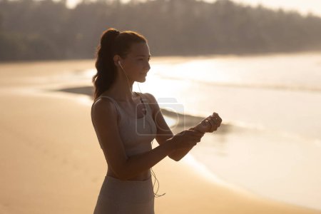 Téléchargez les photos : Silhouette de femme sportive en bonne santé en vêtements de fitness mesure pouls à la main sur la plage le matin. Photo de haute qualité - en image libre de droit