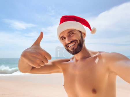 Photo for Happy bearded beach man on Christmas travel holidays taking selfie picture with smartphone wearing santa hat showing thumb up during his winter vacation. Young adult slim guy in front of the seashore - Royalty Free Image