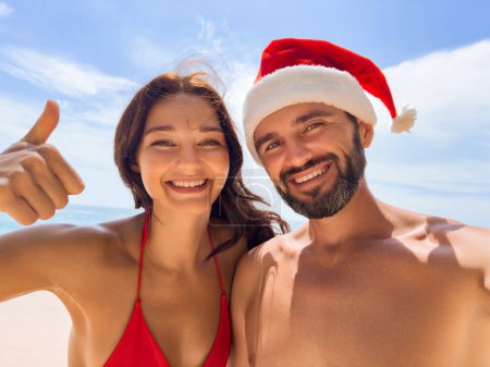 Photo for Christmas couple selfie picture on beach vacation. Happy young adults smiling at camera taking self-portrait with thumb up wearing Santa hat. Multiracial Caucasian and Asian people. High quality photo - Royalty Free Image