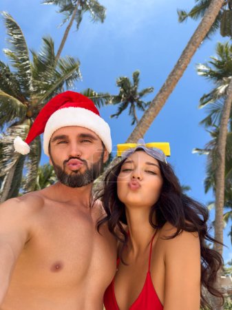 Téléchargez les photos : Couple de Noël selfie photo pendant les vacances à la plage pendant les vacances d'hiver contre les palmiers. Joyeux jeunes adultes souriant à la caméra prenant autoportrait envoyer baiser d'air, portant le chapeau de Père Noël. Multiracial - en image libre de droit
