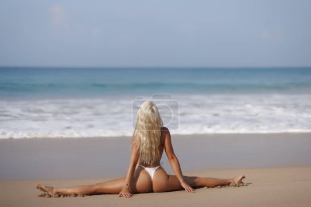 Foto de Hermosa modelo de mujer caucásica bañada por el sol con el pelo rubio en un traje de baño elástico blanco haciendo yoga pierna partida Vista trasera de la chica fitness sexy con figura deportiva perfecta posando en una playa de arena de verano en - Imagen libre de derechos