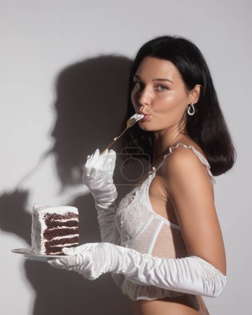 Photo for Stunning fashionable luxurious girl with bright evening makeup dressed in white lacy lingerie tasting a slice of birthday cake, looking at the camera. Fashion lingerie brand anniversary or promotion - Royalty Free Image