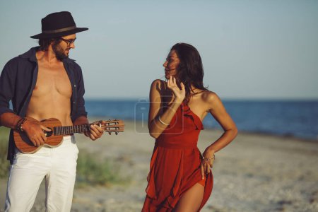 Téléchargez les photos : Couple heureux en amour portrait sensuel. Homme caucasien jouant ukulélé sérénade et fille en robe rouge dansant sur la plage au coucher du soleil. Un homme beau barbu et une femme magnifique ayant tendre romantique - en image libre de droit