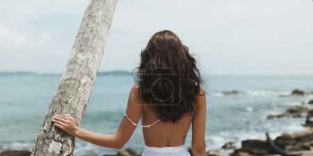 Photo for Vacation of beach fashion multiracial woman model with perfect slim tanned body in stylish white modern swimwear at a palm tree. Rear view of the perfect girl in full-length - Royalty Free Image