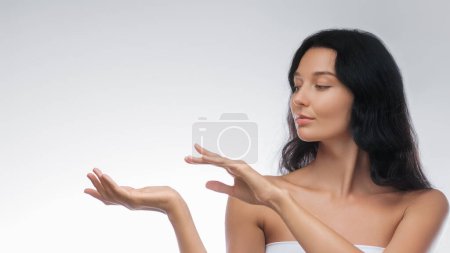 A graceful woman with long black hair presents an invisible product with poised hands against a neutral background. Perfect for product advertisements, beauty, and skincare promotions. 