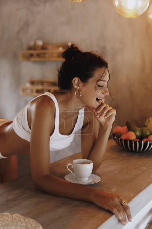 Eine braun gebrannte Frau in minimalistischer Kleidung genießt ihren Morgenkaffee in einer stilvollen Küche, die von warmem Licht erhellt wird. Dieses hochwertige Bild ist perfekt für die Förderung von Konzepten des modernen Lebensstils, Morgen