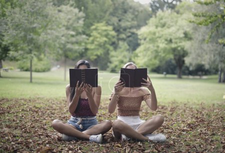 Foto de Dos mujeres jóvenes con rostros cubiertos por dos libros, sentadas en un parque. - Imagen libre de derechos