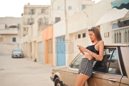 Foto de Retrato de una joven apoyada en un coche de época con un smartphone en la mano - Imagen libre de derechos