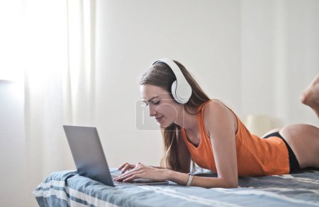 Photo for Young woman lying on the bed uses a computer and listens to music - Royalty Free Image