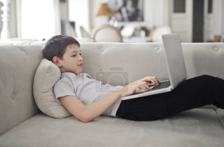 Foto de Niño utiliza un ordenador portátil tumbado en el sofá en casa - Imagen libre de derechos