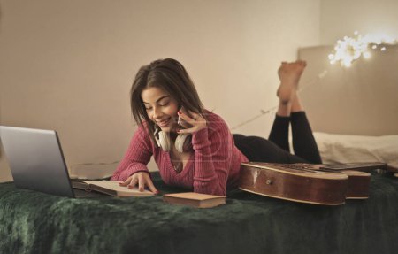 Foto de Mujer joven utiliza una computadora y un teléfono inteligente tumbado en la cama - Imagen libre de derechos