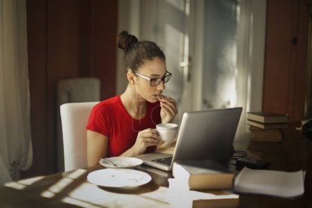 Foto de Mujer joven utiliza una computadora en casa mientras come galletas con té - Imagen libre de derechos