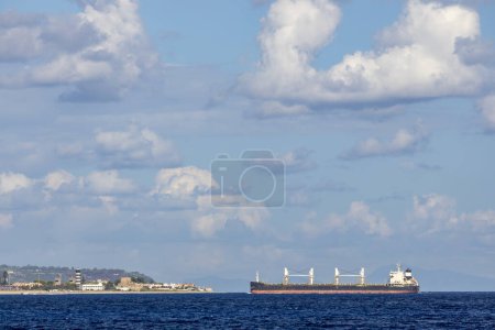 Photo for Cargo ship nearby Capo Peloro Lighthouse in Punta del Faro on the Strait of Messina, most north eastern promontory of Sicily, Italy - Royalty Free Image